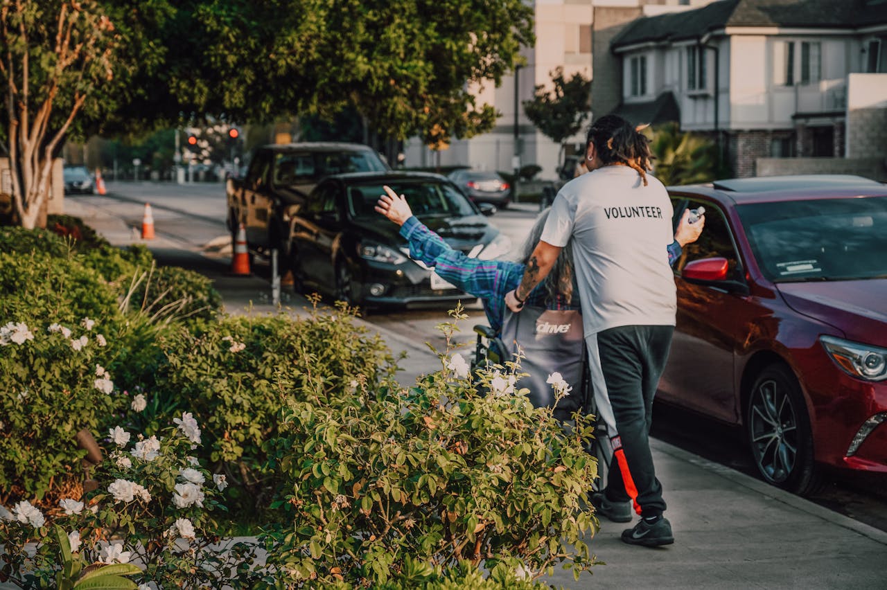 services-02 Volunteer assisting a person in a wheelchair on a city street surrounded by greenery and parked cars.