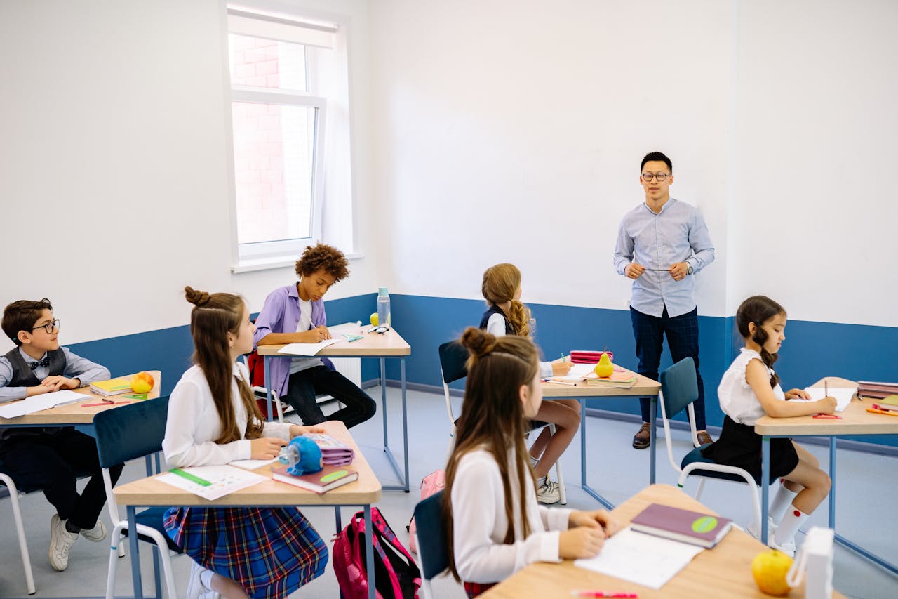 hero-img-01 Teacher instructing a diverse group of elementary students in a modern classroom setting.